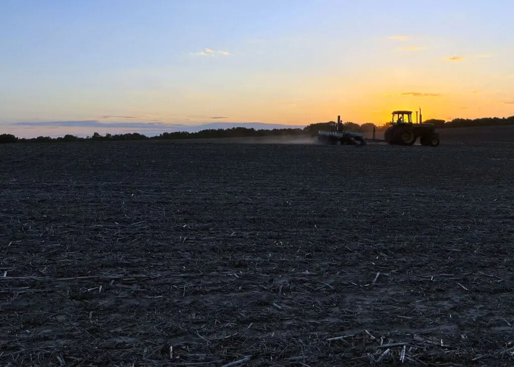 A tractor sits in a field while the sun rises in the background. 
